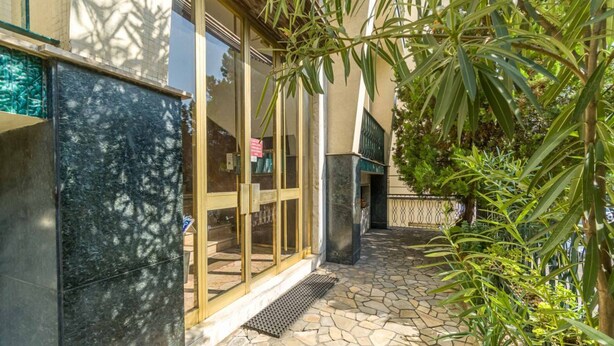 Une entrée d’immeuble avec un chemin pavé de pierres, des portes en verre et de la verdure environnante.