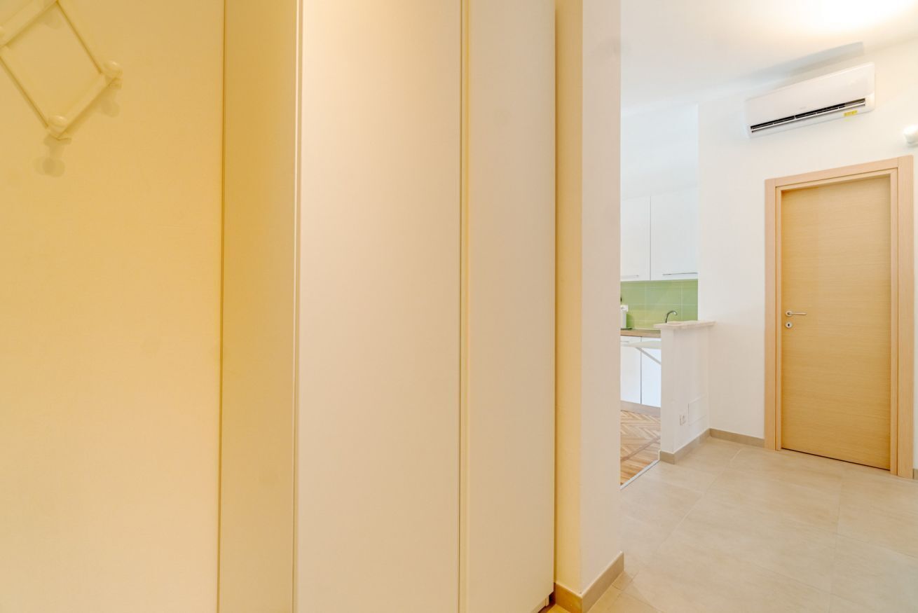 Un couloir avec une porte en bois, un mur blanc et un aperçu d’une cuisine dont le plan de travail est recouvert d’un revêtement vert.
