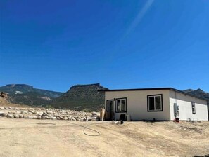 Il s’agit d’un bâtiment moderne doté d’un toit plat, de grandes fenêtres et d’une porte. Il est situé sur un terrain aride et rocheux, avec des montagnes en arrière-plan.