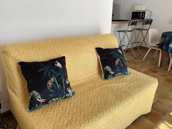 A yellow quilted sofa with two decorative pillows featuring jungle animals, positioned in a room with a tiled floor and a small dining area in the background.