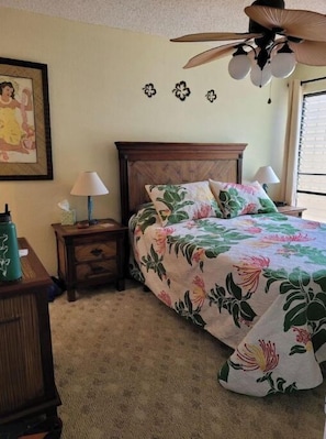 Un dormitorio con cabecera de madera, colcha estampada con motivos florales, dos lámparas de noche, ventilador de techo y un cuadro en la pared.