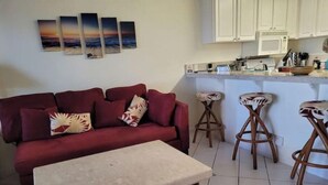 Sala de estar con un sofá rojo, una mesita y una zona de cocina con armarios blancos y un taburete de bar.