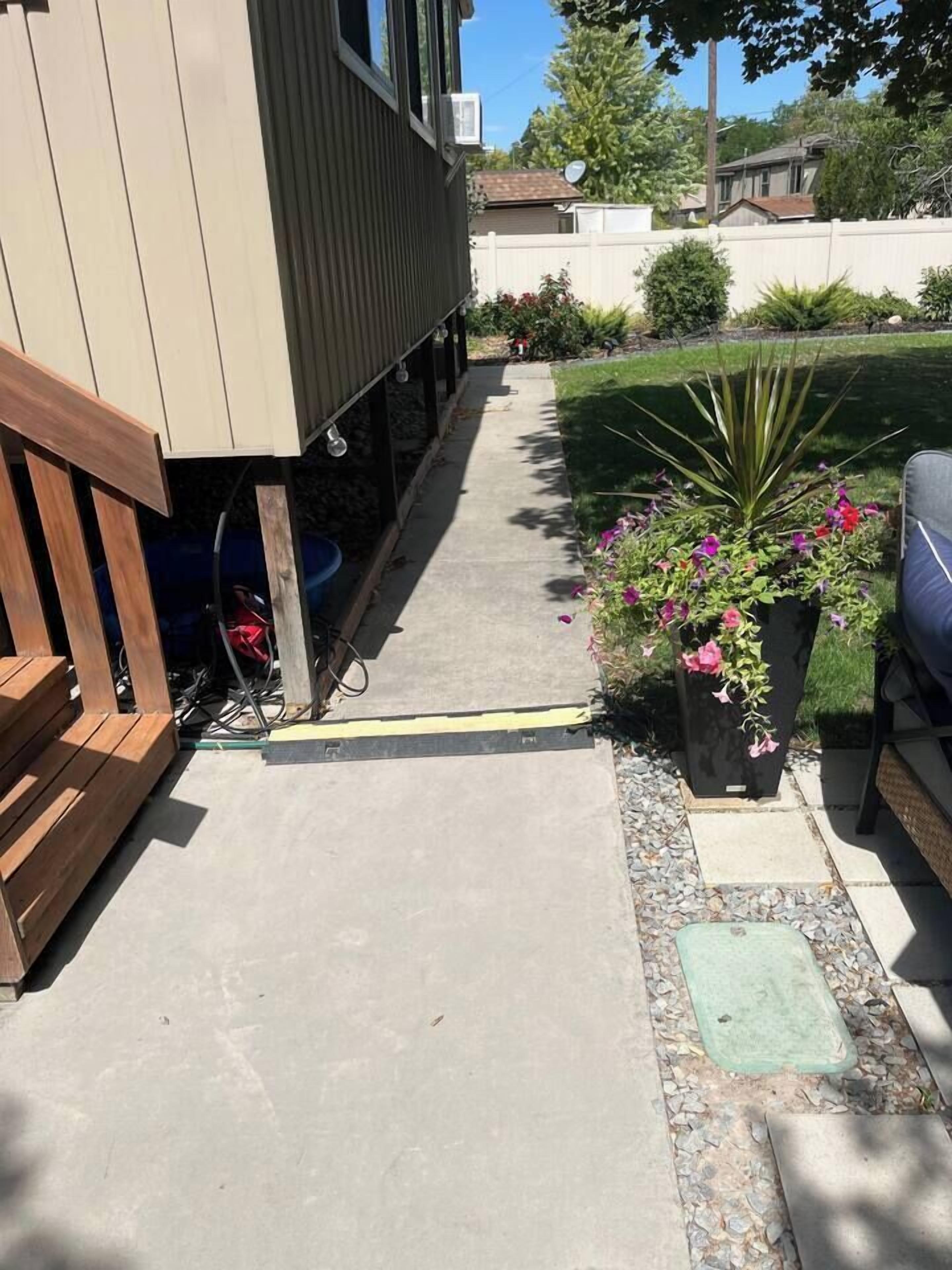 A concrete pathway leading to a house with a wooden deck and a potted plant with pink flowers.