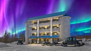 A multi-story hotel with a blue roof and a covered entrance area, surrounded by snow and parked vehicles. - Hatcher Pass Castle|All Inclusive|Mountain View (Willow)