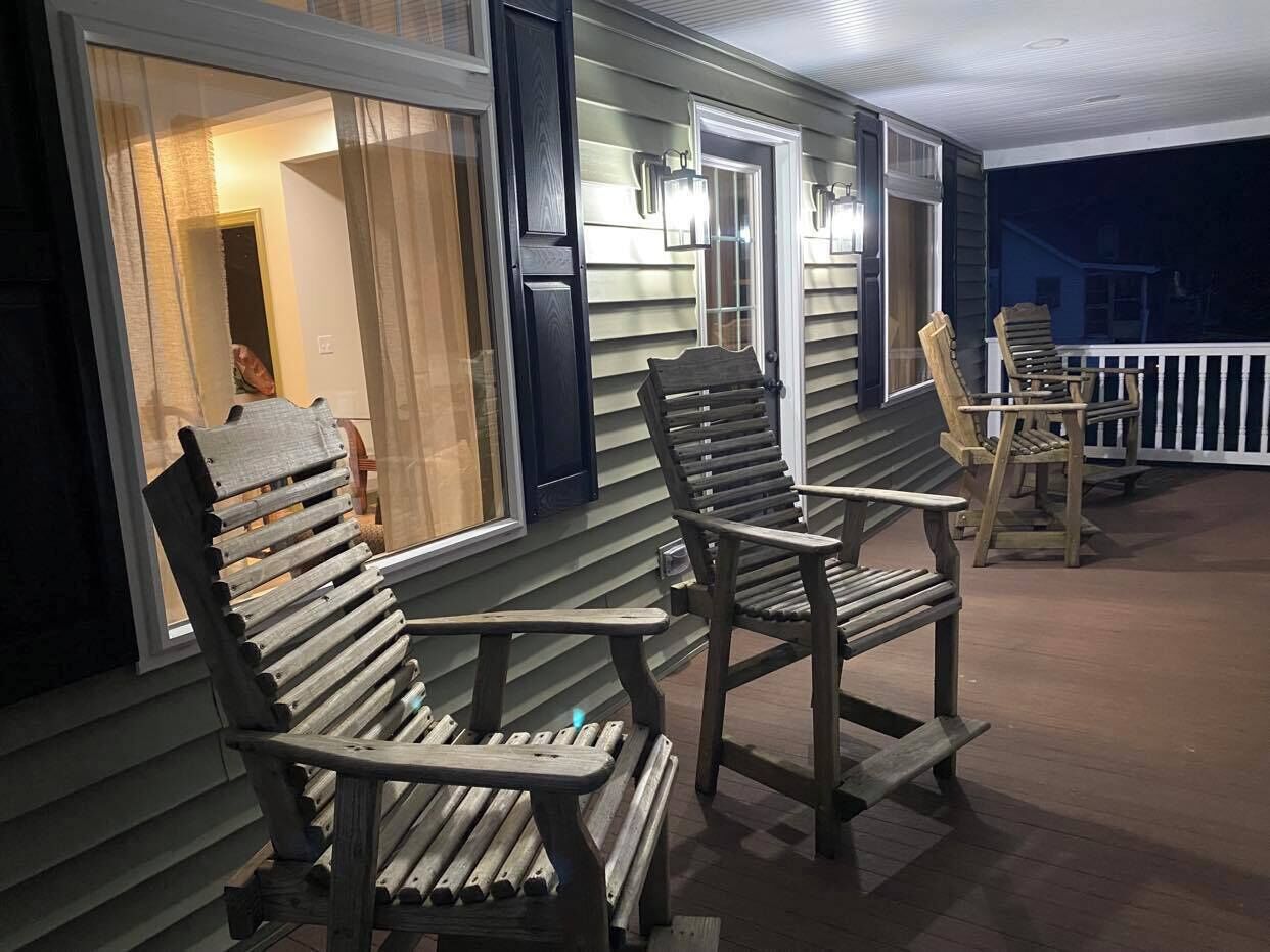 Een veranda met twee houten Adirondack-stoelen en een klein tafeltje, een deur en een raam met uitzicht op het interieur.