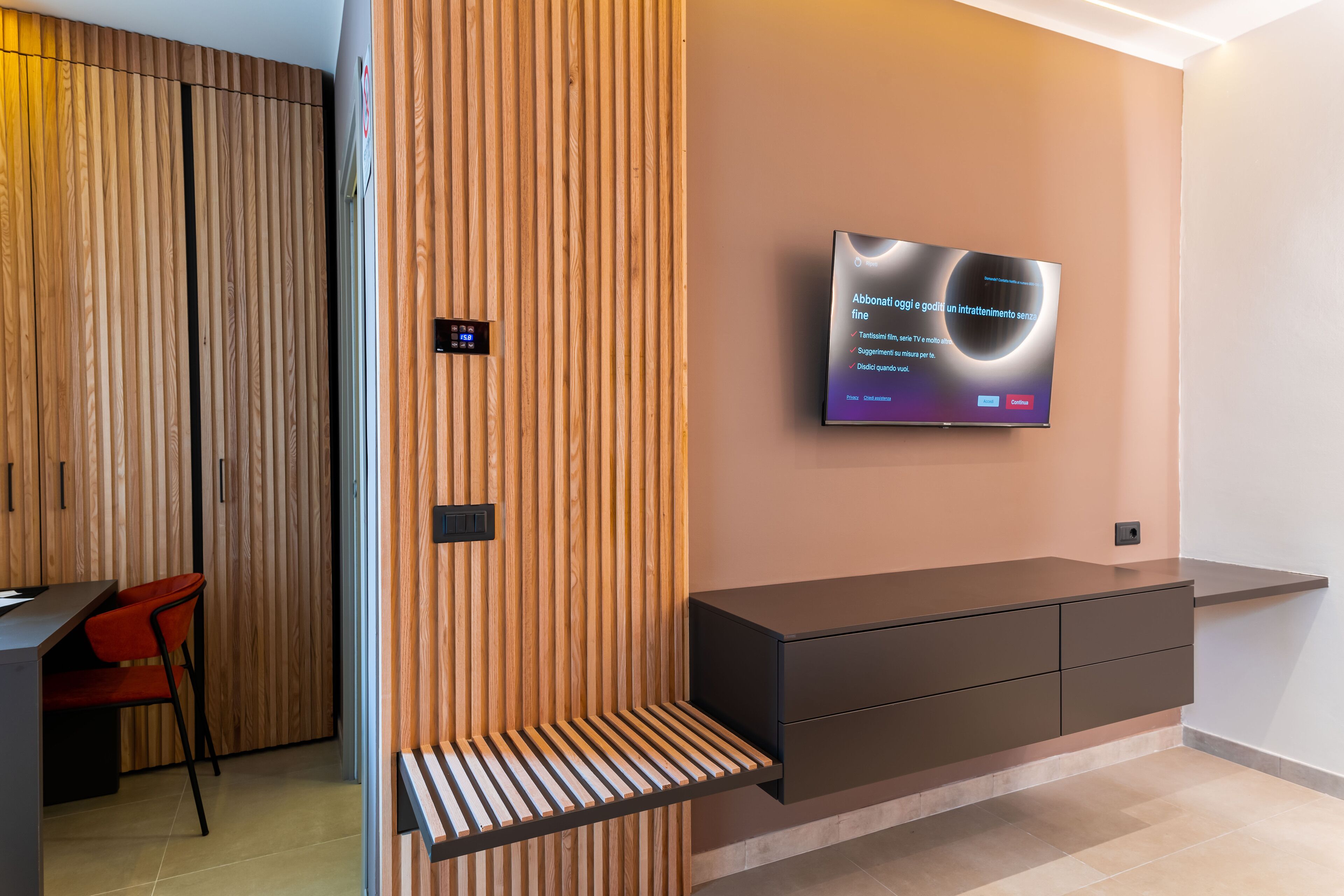 Ein modernes Interieur mit einer Holzpaneelwand, einem Wand-Fernseher, einem dunklen Schreibtisch mit einem roten Stuhl und einer integrierten Bank.