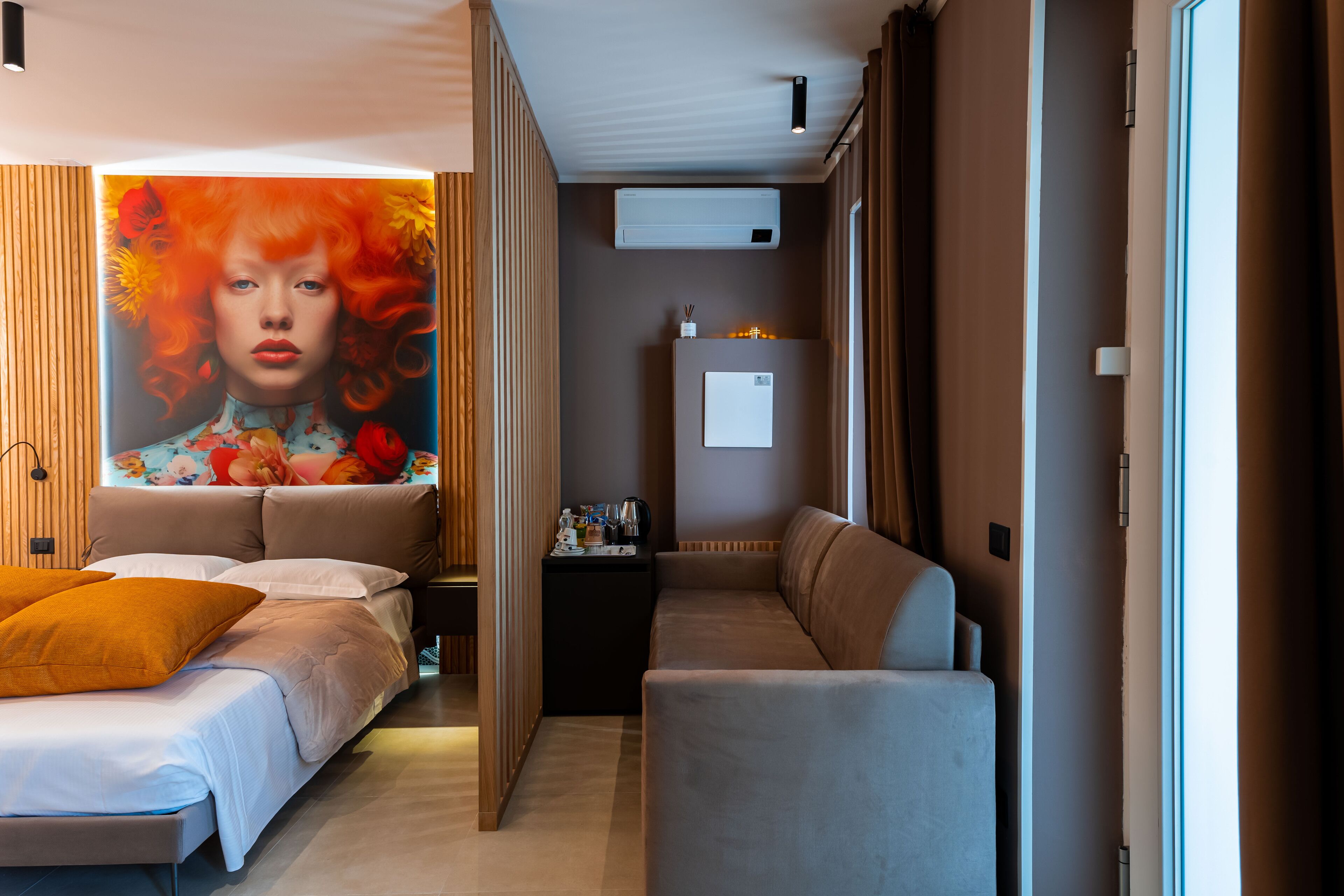 A modern hotel room with a bed, a sofa, a small table, and an air conditioning unit.