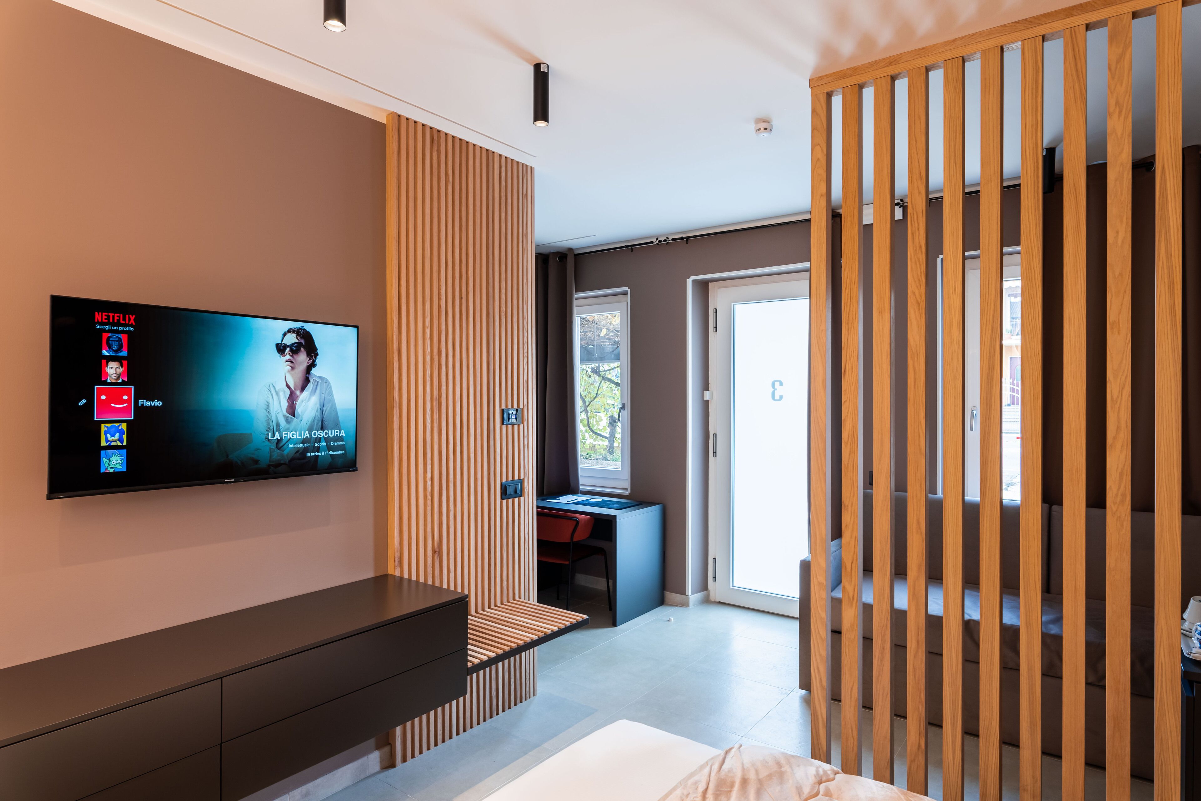 Un salon moderne avec un téléviseur à écran plat fixé au mur, un séparateur en bois, un bureau avec une chaise et un escalier menant à un autre niveau.