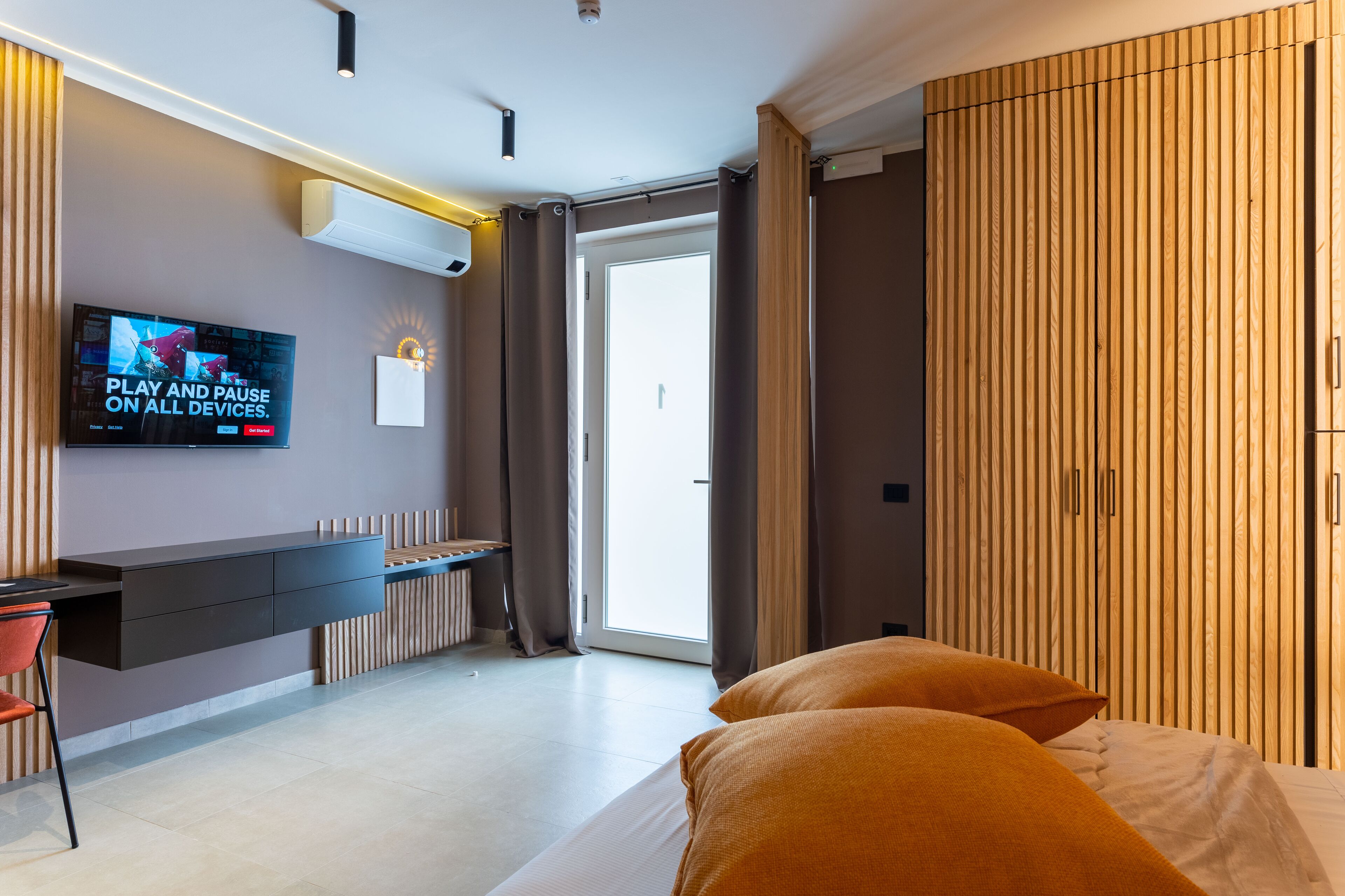 Ein modernes Hotelzimmer mit flachbildfernseher an der Wand, einem Schreibtisch mit Stuhl, einem Bett mit zwei Kissen und einem Kleiderschrank mit vertikalen Lamellen.
