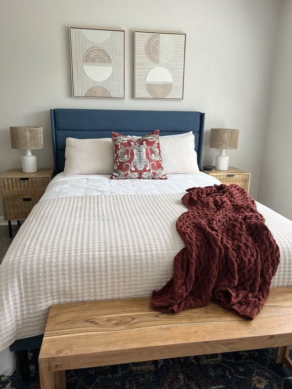 A neatly made bed with a white quilt, a red patterned pillow, and a dark red blanket on a wooden bench. There are two bedside tables with lamps and a headboard. - All Things Auburn, Walkable to Campus and University Related Activities! (Auburn)
