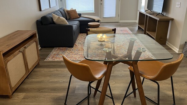 Ein Wohnzimmer mit Esstisch, zwei Stühlen, einer Couch, einem Fernseher und einer Holzkommode.