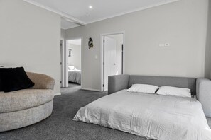 A bedroom with a bed, a grey sofa, and a curved armchair. There is a door leading to another room.
