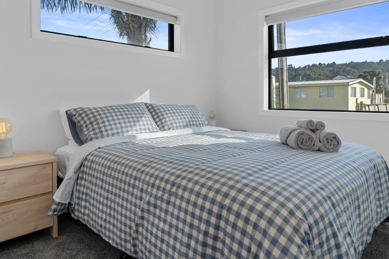 A bedroom with a checkered bedspread, a bedside lamp, and a view of a house through a window.