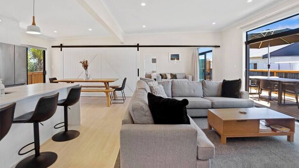 A modern living room with a grey sofa, a wooden coffee table, and a dining area with a white table and black chairs. - Leisure on Leo (Waihi Beach)