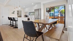 A modern kitchen with white cabinets, a stainless steel refrigerator, and a wooden dining table with a bench. There are bar stools and pendant lights above the counter. - Leisure on Leo (Waihi Beach)
