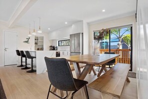 Uma cozinha moderna com armários brancos, geladeira de aço inoxidável e uma mesa de jantar de madeira com um banco. Há banquetas e luminárias pendentes sobre o balcão.