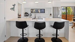 A modern kitchen with a central island, three black bar stools, a flat-screen TV, and a stainless steel refrigerator. - Leisure on Leo (Waihi Beach)