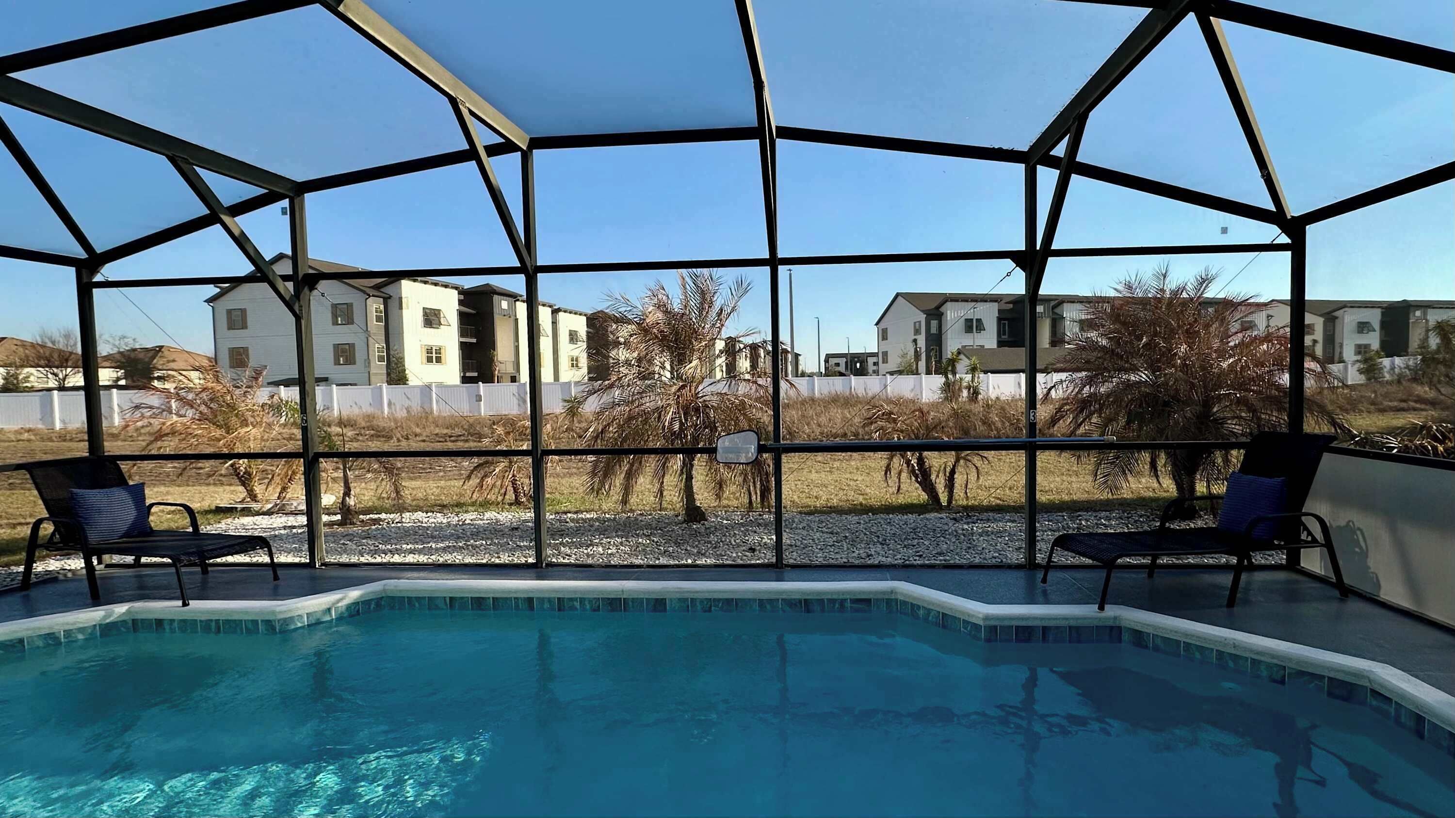 Une piscine entourée de parois en verre, deux chaises longues et une vue sur des immeubles résidentiels.