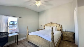 Um quarto com cama, escrivaninha, cadeira, mesa de cabeceira e ventoinha no teto.