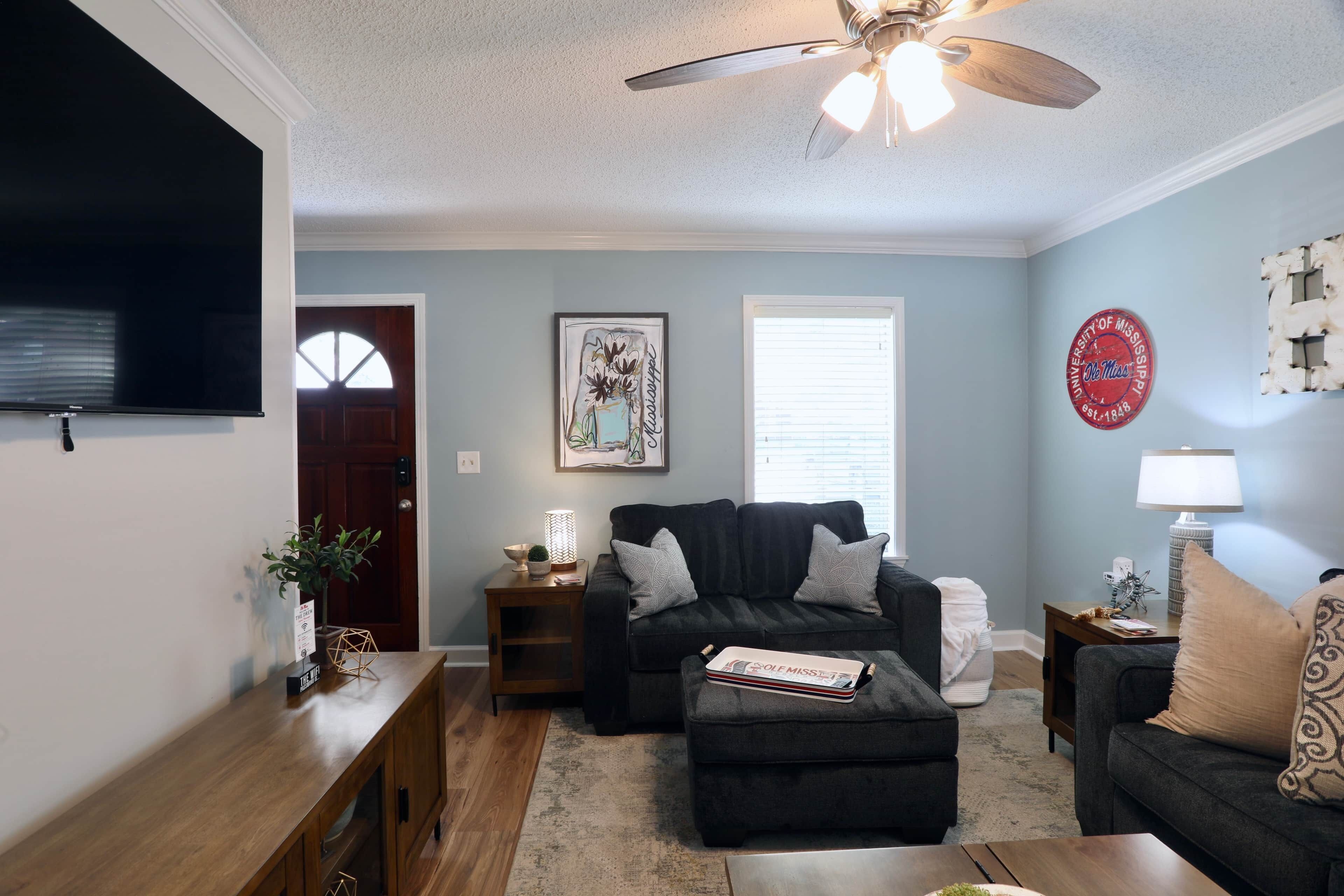 A living room with a dark grey sofa, a wooden coffee table, a ceiling fan, and a wall-mounted TV.