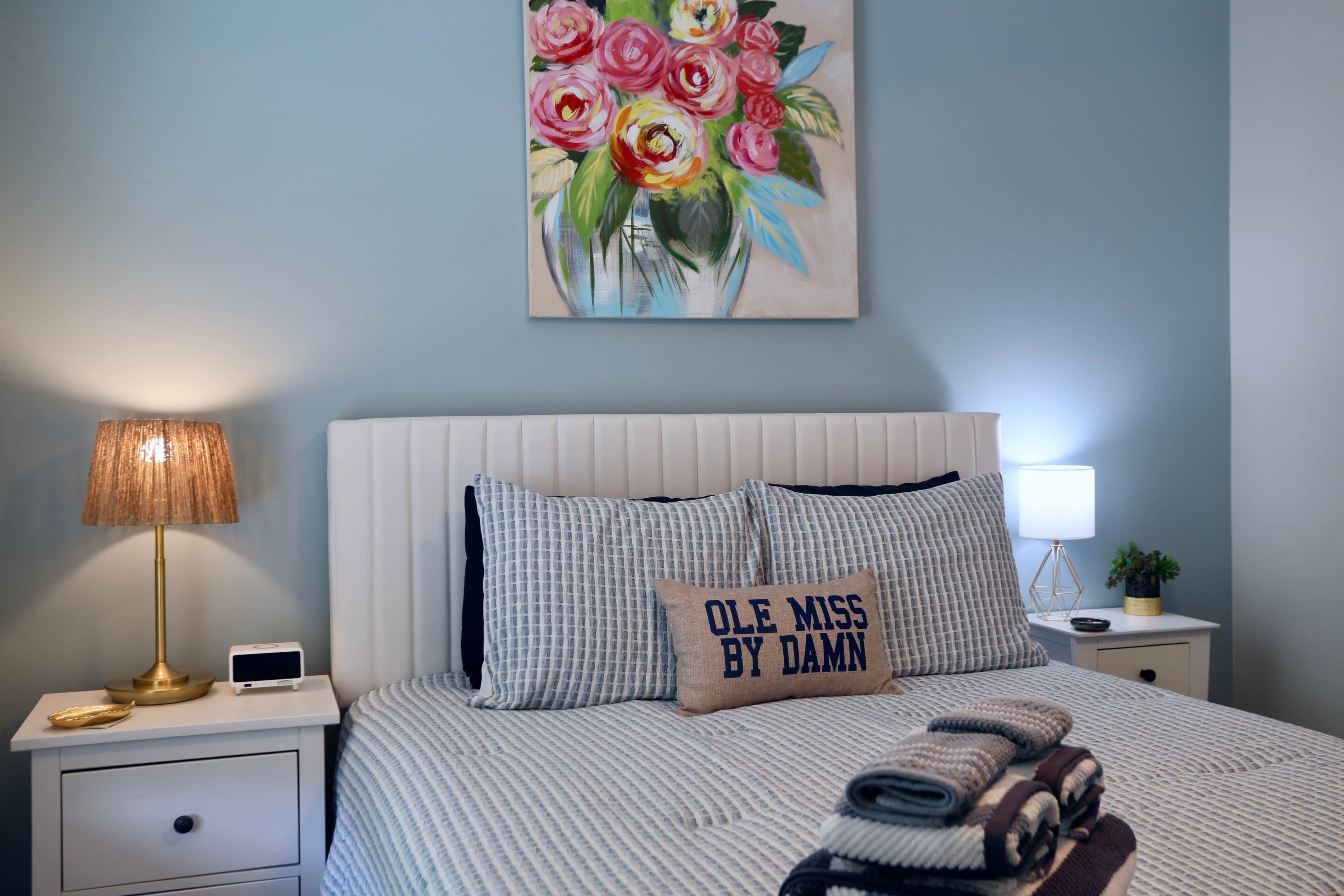 Een slaapkamer met een bed, een bedlamp, een nachtkastje, een wandklok en een bloemenstilleven.