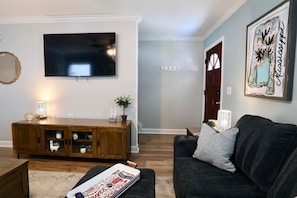 Una sala de estar con una televisión de pantalla plana, un sofá de color oscuro, un mueble de madera y una mesita con objetos decorativos.