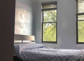 A bedroom with a bed, a headboard, and two windows with blinds.