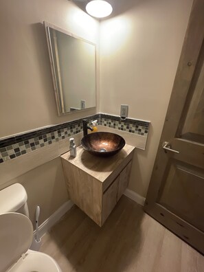 A bathroom with a wooden vanity, a copper vessel sink, a mirror, and a toilet.