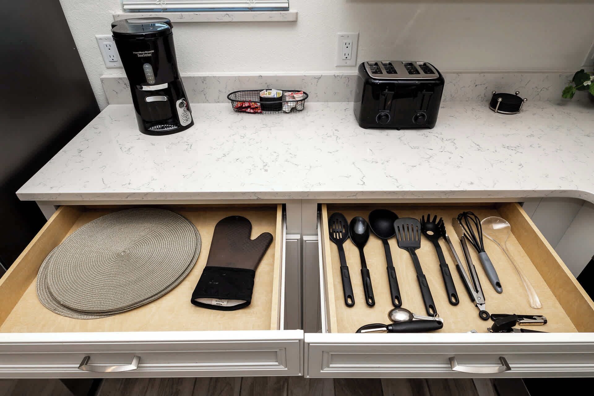 A kitchen counter with a coffee maker, toaster, and a basket of snacks. Below, a drawer contains kitchen utensils and a cutting board.