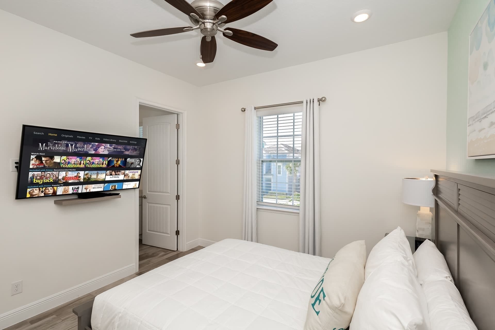 A bedroom with a bed, a ceiling fan, a TV mounted on the wall, and a window with blinds.