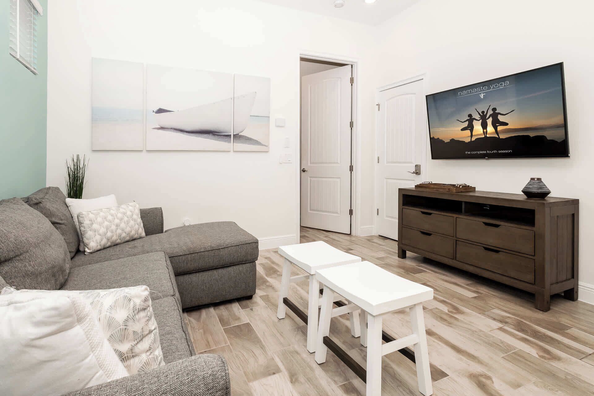 A living room with a grey sectional sofa, a white coffee table, a wooden TV stand with a flat-screen TV, and a door leading to another room.