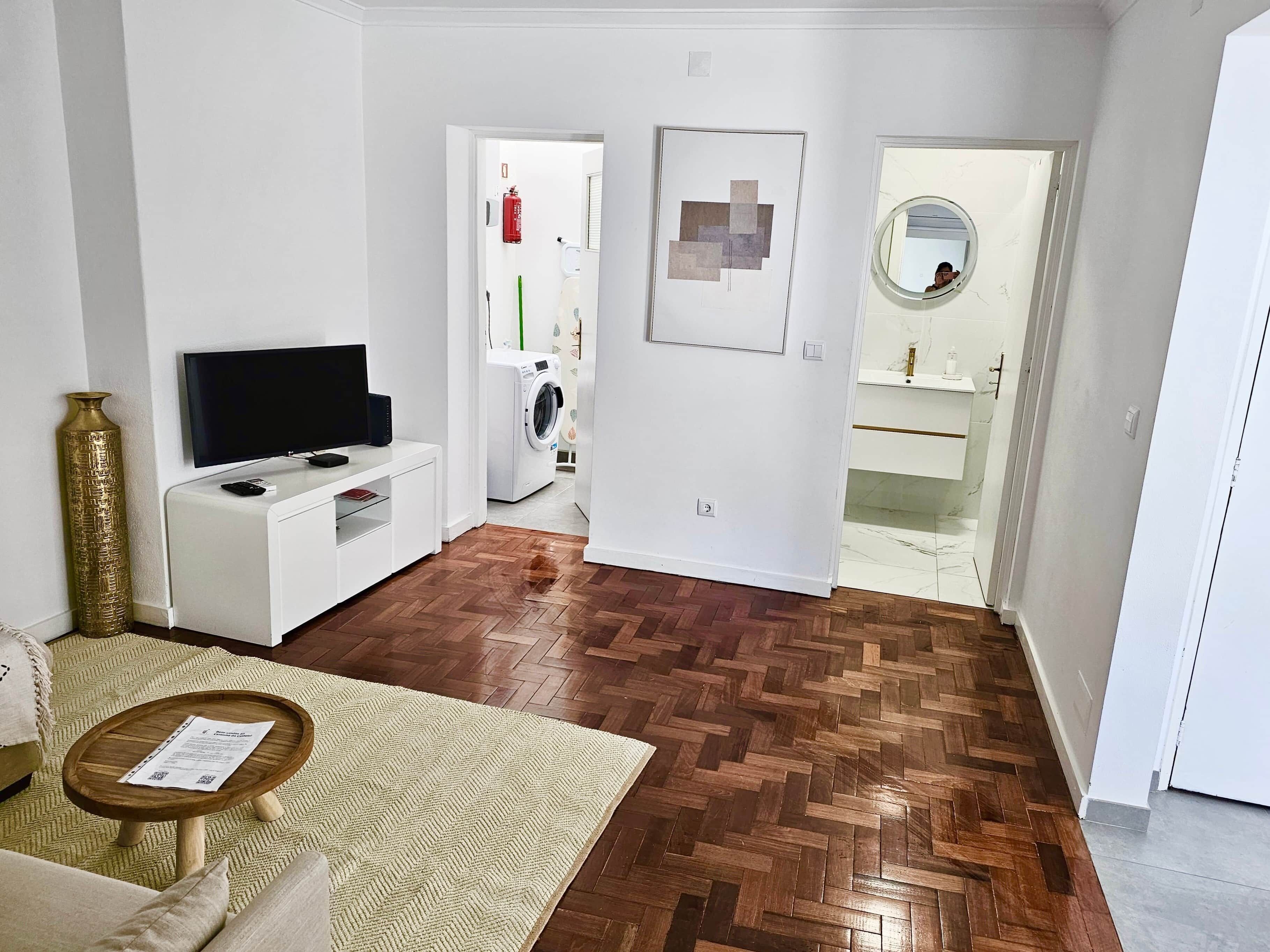 Ein Wohnzimmer mit Fernseher auf einem weißen Schrank, Holzboden im Fischgrätmuster, einem kleinen Holztisch mit einem Dokument und einer Couch.