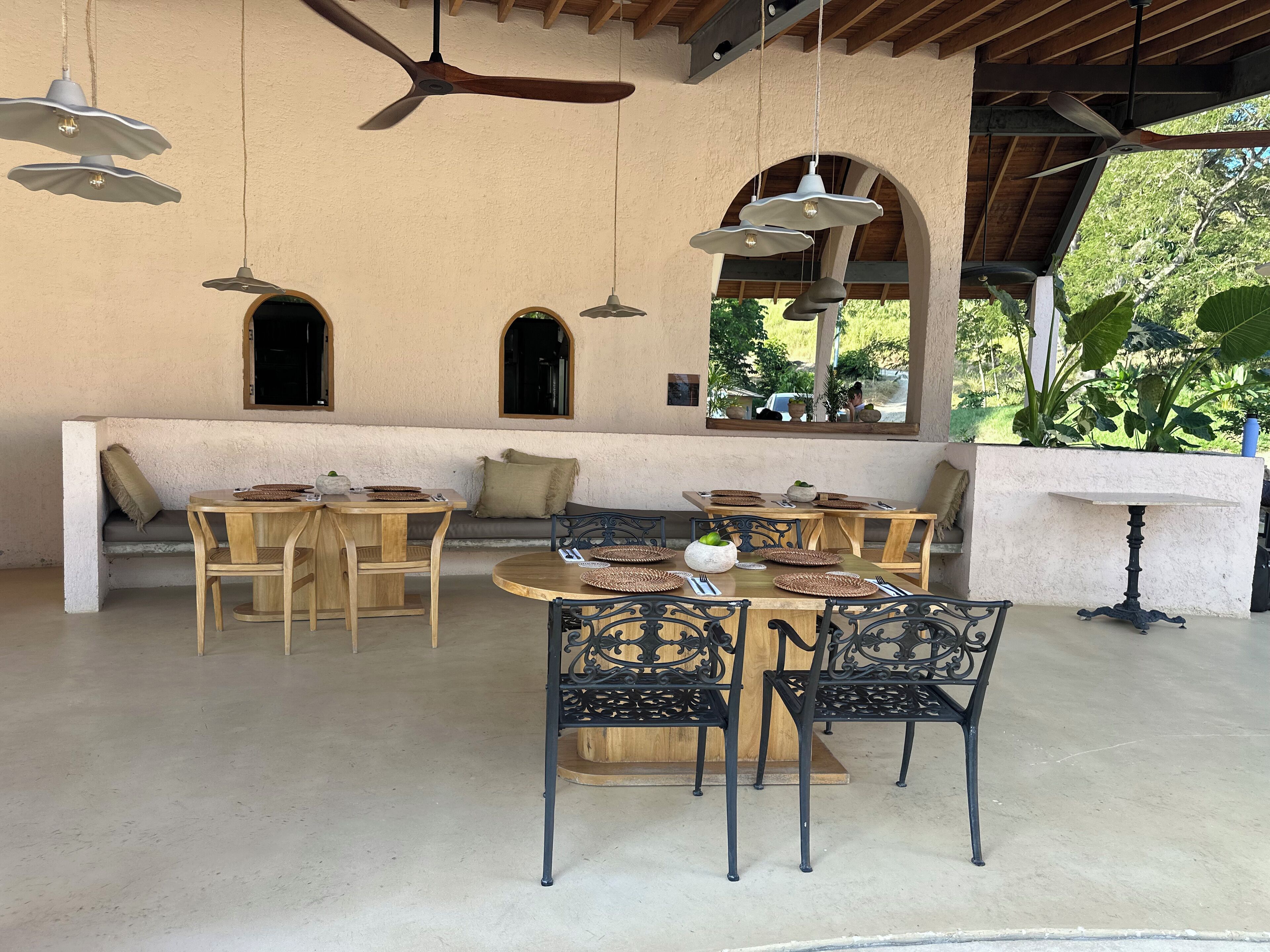 Un espace extérieur abrité, aménagé pour les repas, avec des tables et des chaises en bois, un banc et des ventilateurs de plafond.