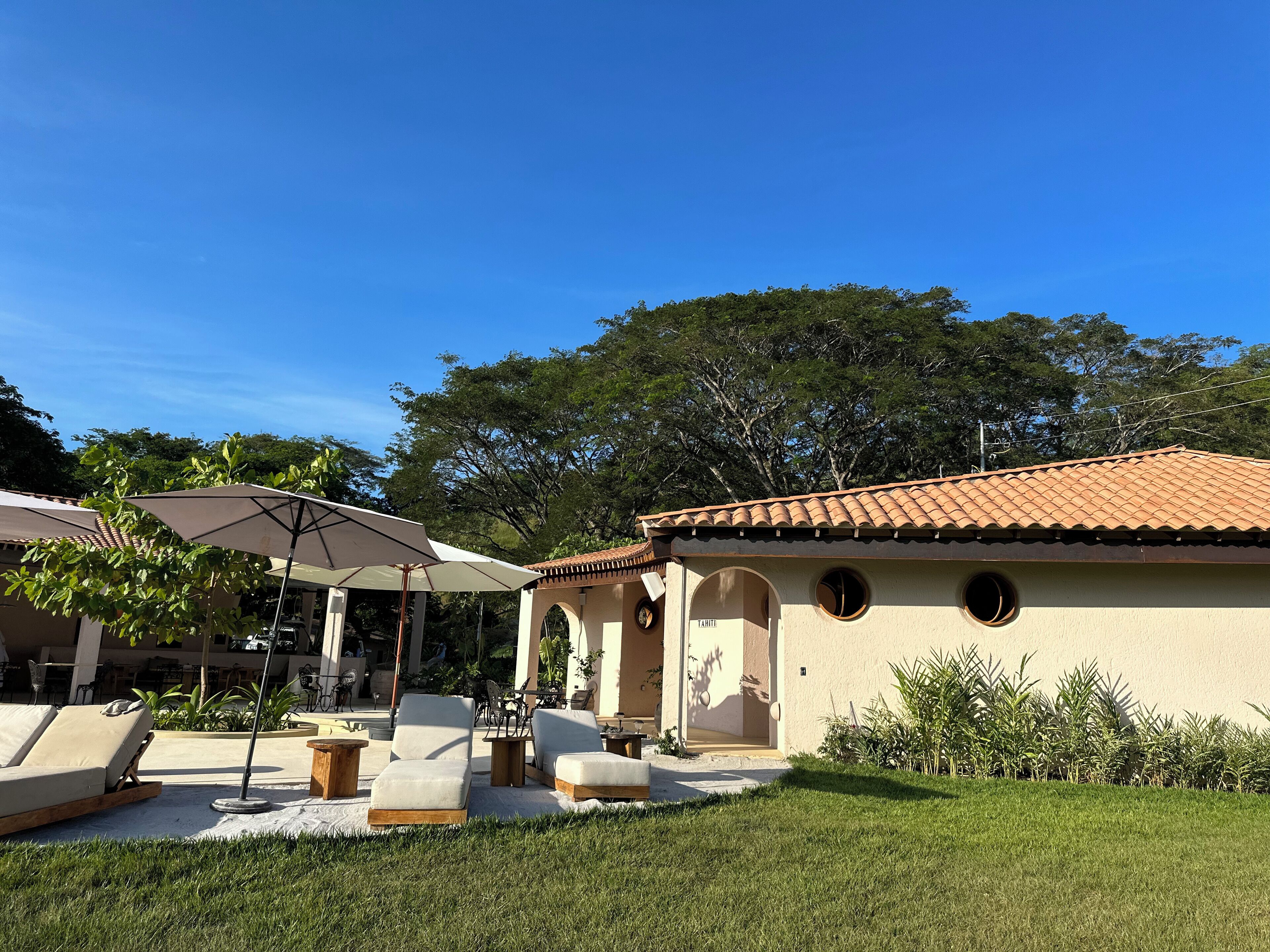 A sunny outdoor seating area with lounge chairs and umbrellas, a building with a tiled roof, and a well-maintained lawn.