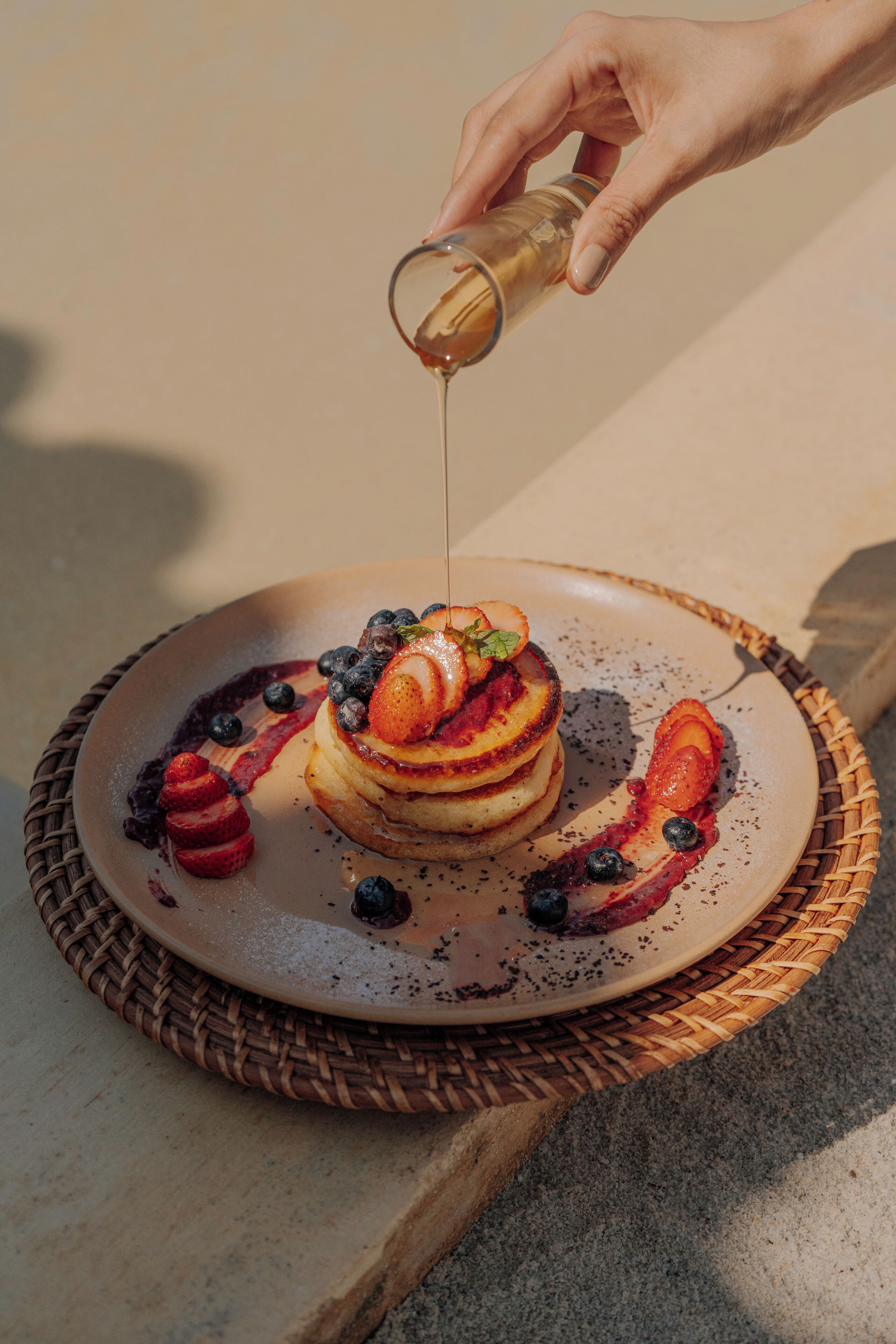 A hand pouring syrup over a stack of pancakes on a plate.