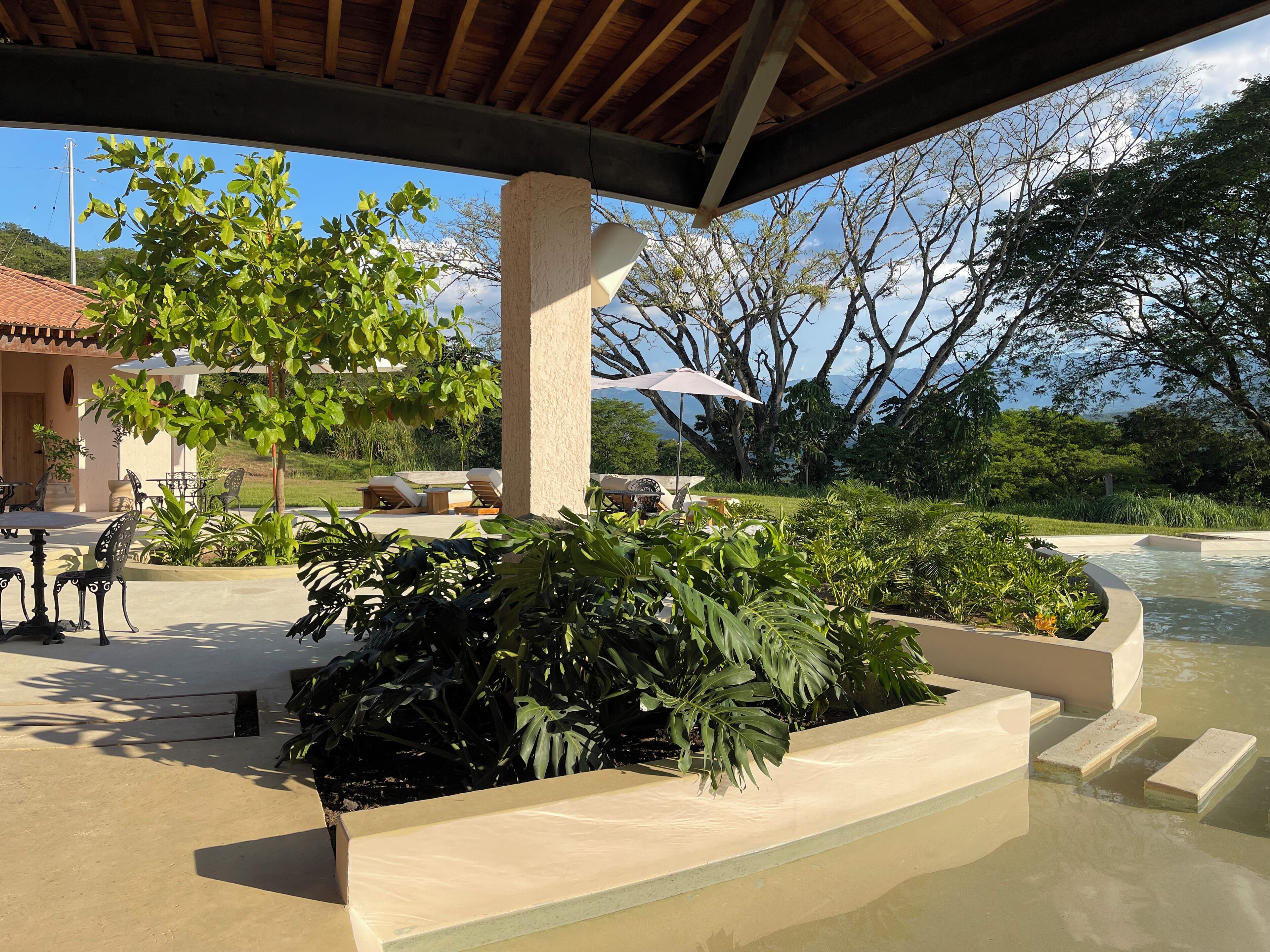 An outdoor area with a covered seating area, a pool, and lush greenery.