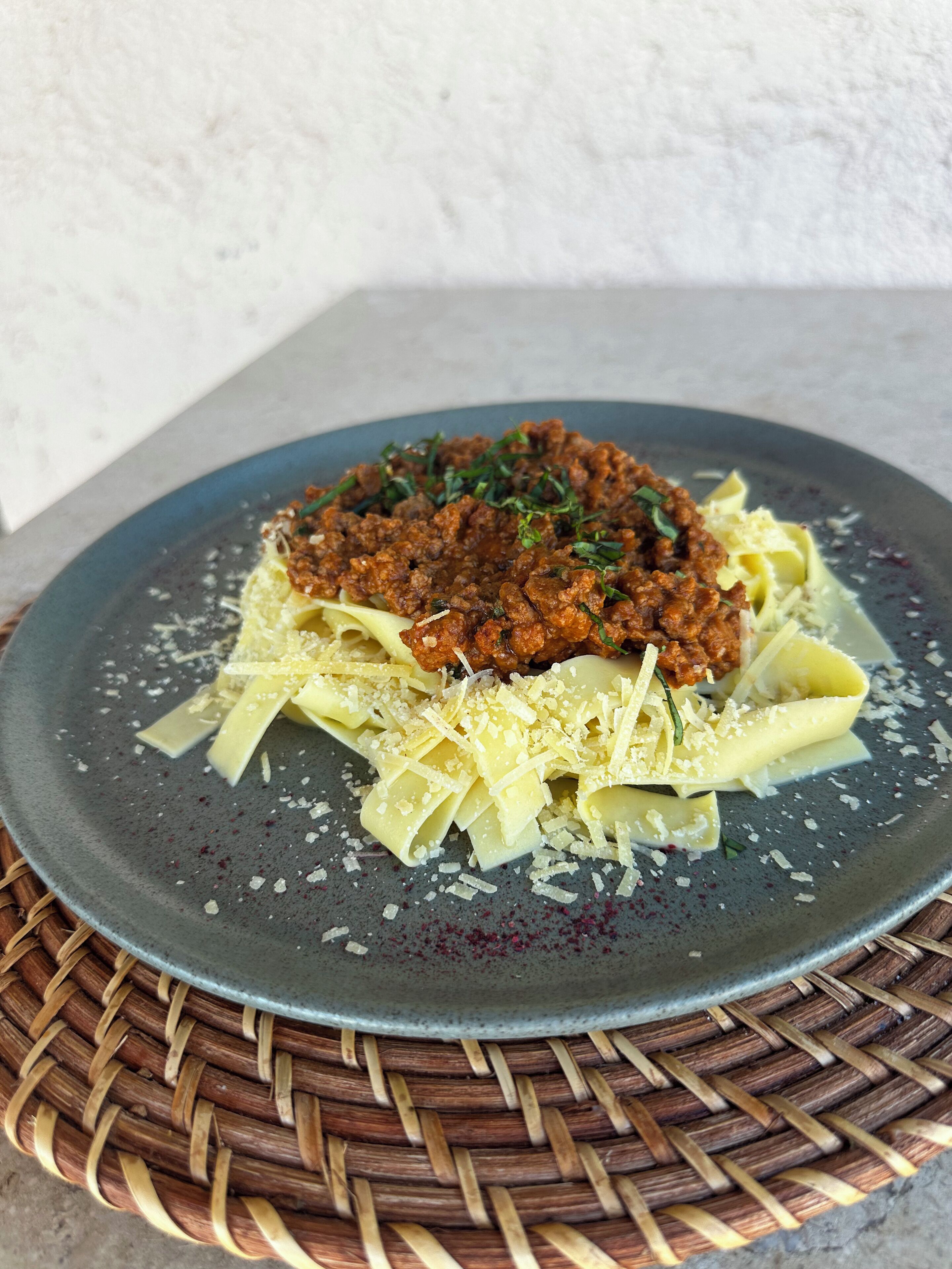 A plate of pasta with a meat topping, garnished with herbs, on a dark plate with grated cheese.
