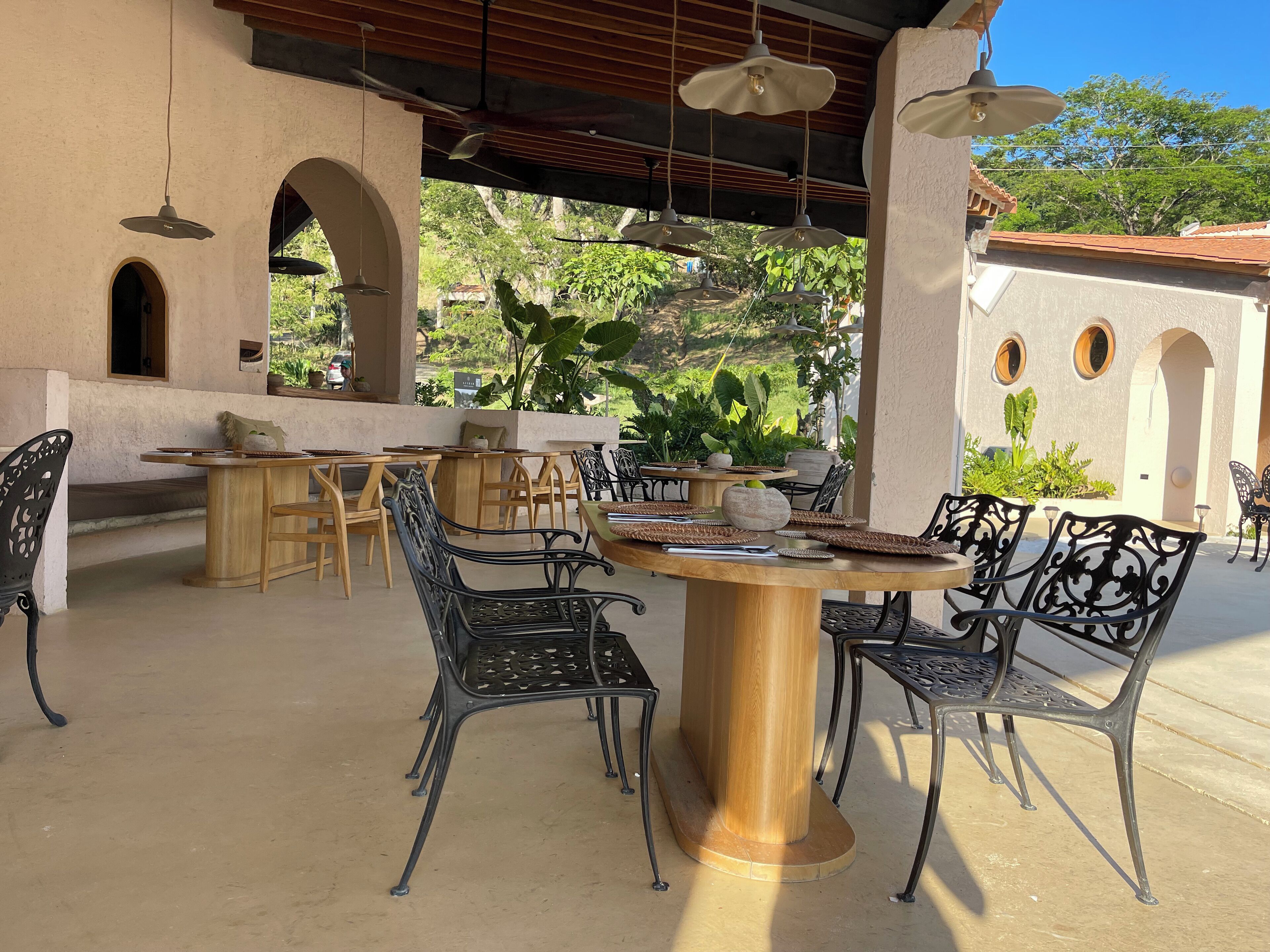 An outdoor patio with round tables and chairs, surrounded by plants and architectural arches.