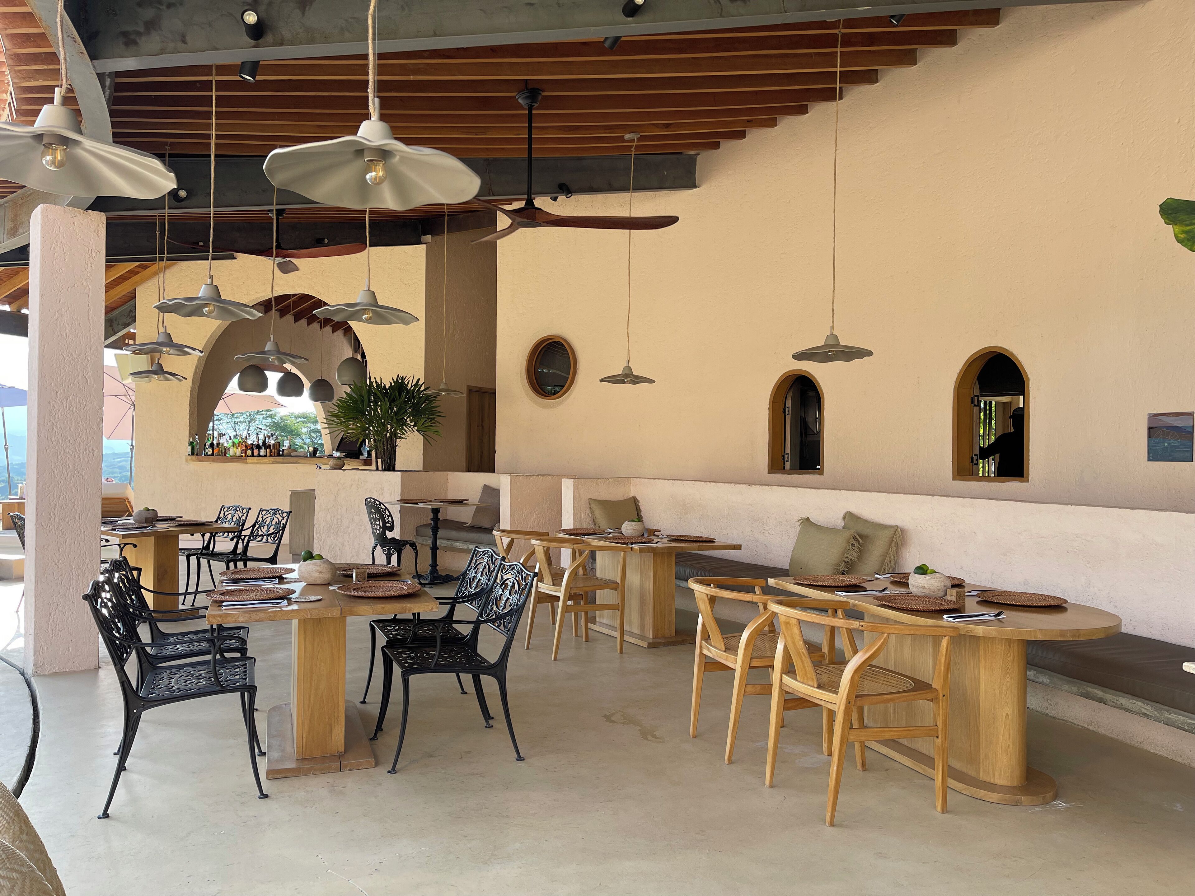 Un espacio de comedor al aire libre con varias mesas y sillas, lámparas colgantes y vistas al paisaje.