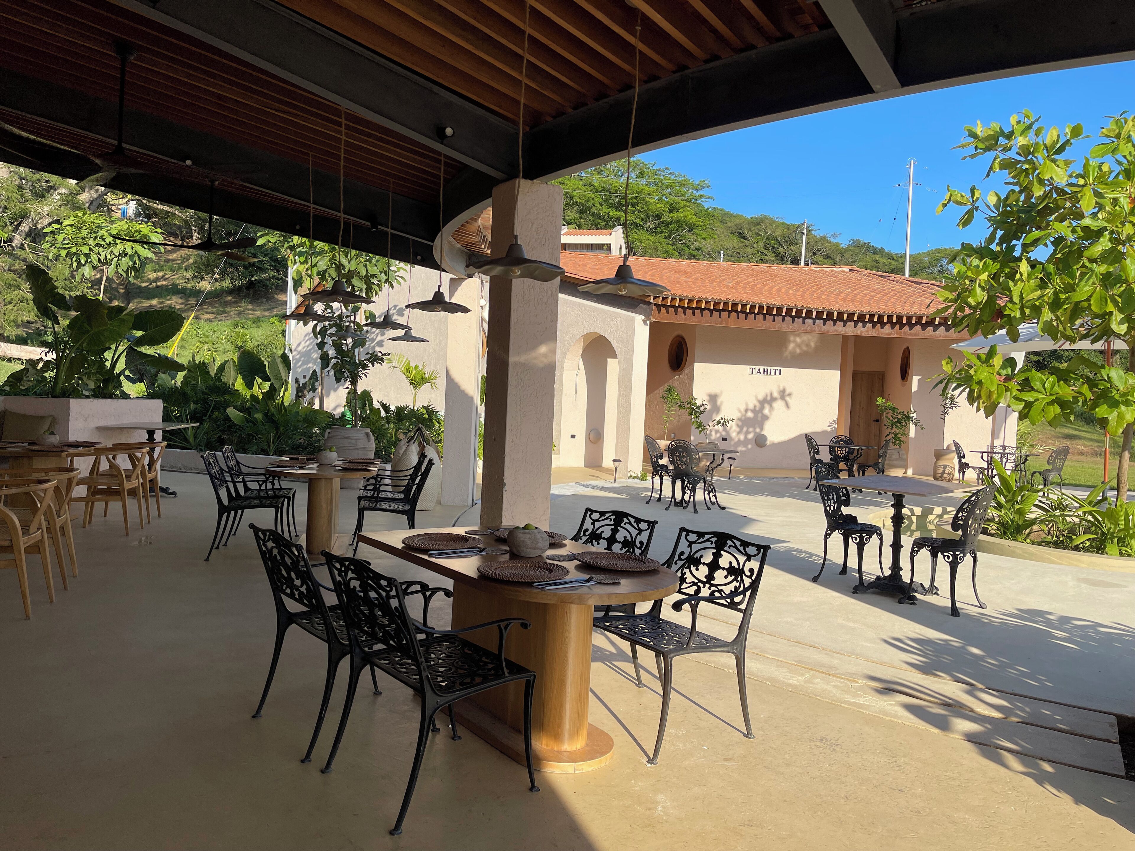 Un patio al aire libre con mesas y sillas, rodeado de follaje y un edificio con techo de tejas.