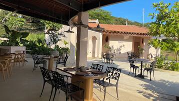 Un patio al aire libre con mesas y sillas, rodeado de follaje y un edificio con techo de tejas.