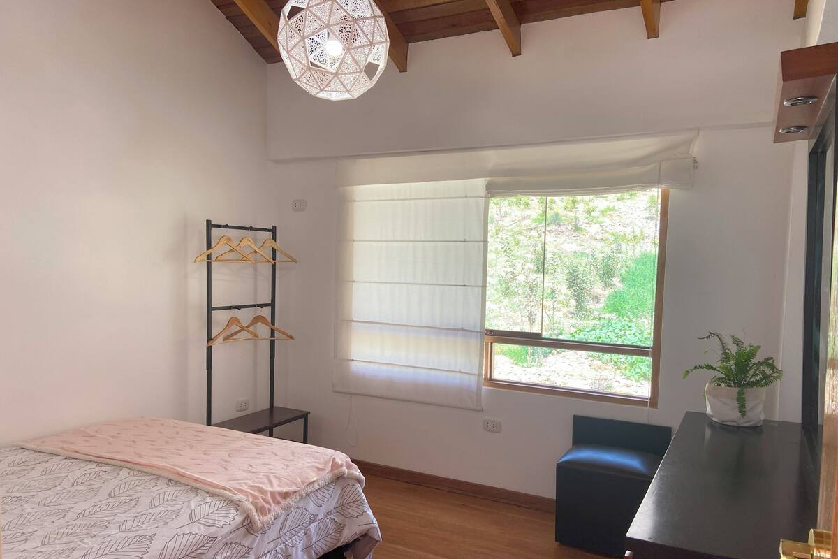 Une chambre avec un lit, une fenêtre avec des stores, un portant à vêtements, une petite table avec une plante et un banc.