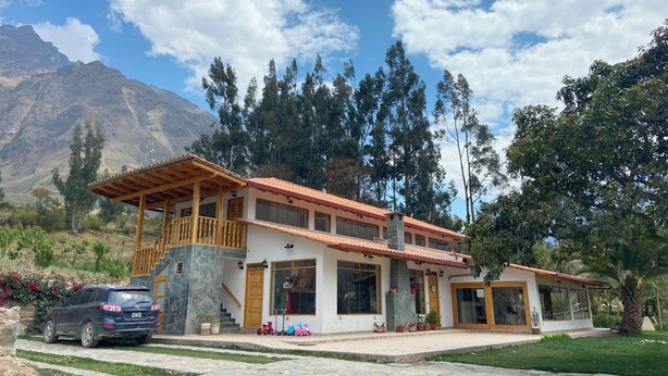 A two-story house with a balcony, a car parked in front, and a mountainous backdrop. - Quinta San Martin (Ollantaytambo)