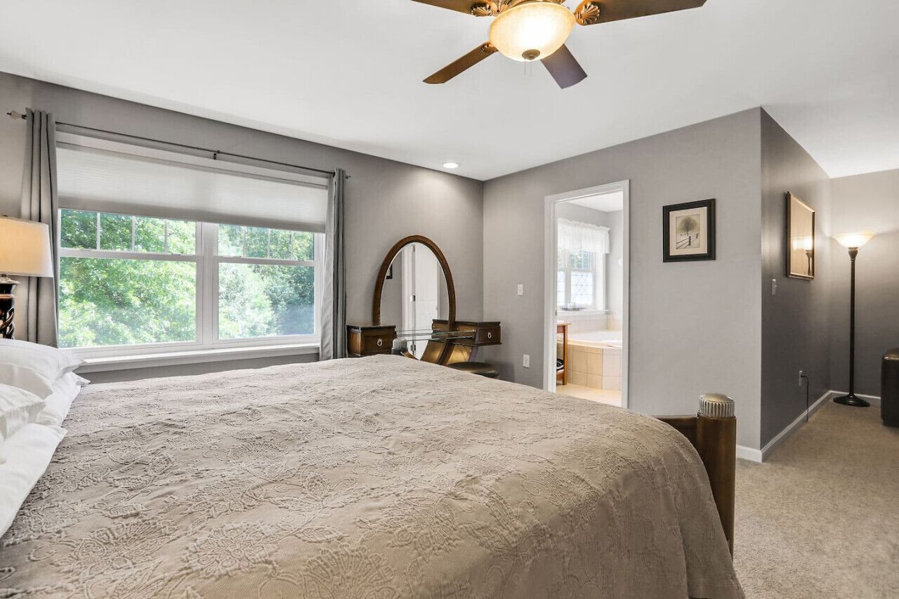 A bedroom with a large bed, a ceiling fan, a window, a mirror, and a door leading to another room.