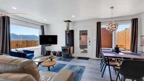 Ein modernes Wohnzimmer mit Fernseher, Kamin, Essbereich und großen Fenstern mit Blick auf eine gebirgige Landschaft.