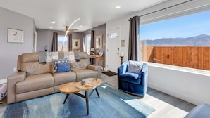 Ein Wohnzimmer mit Sofa, Sessel, Couchtisch und Blick auf Berge durch ein großes Fenster.