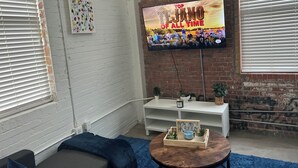 Una sala de estar con un televisor mostrando 'Top LJANO OF ALL TIME', una pared de ladrillo blanco, un sofá azul y una mesa de centro de madera con objetos decorativos.