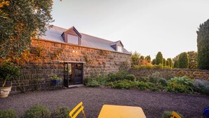 A stone building with a metal roof, a gravel driveway, and a yellow table with chairs in the foreground. - Honeysuckle Barn & Garden (Kyneton)