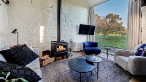 A cozy living room with a stone wall, a fireplace, a comfortable sofa, a blue armchair, and a large window offering a view of the outdoors. - Honeysuckle Barn & Garden (Kyneton)