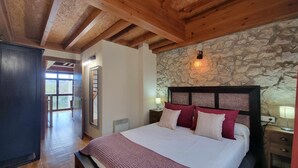 A bedroom with a stone wall, wooden ceiling beams, a bed with multiple pillows, a nightstand, and a window. - Los Gemelos Vacation Home (San Roque Del Acebal)